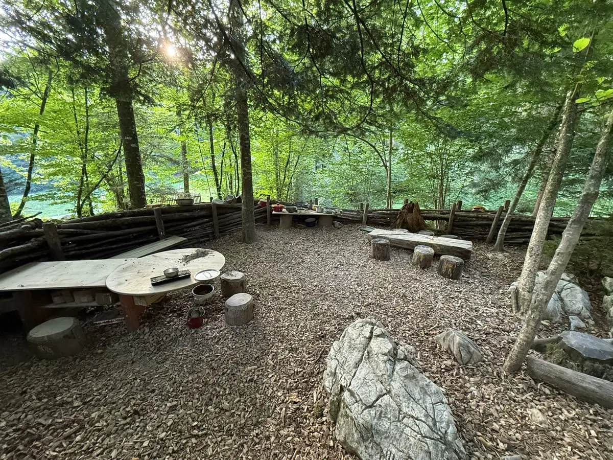 Holztische des Brotzeitplatzes mit Palisadenzaun im Wald, von der Abendsonne beleuchtet.
