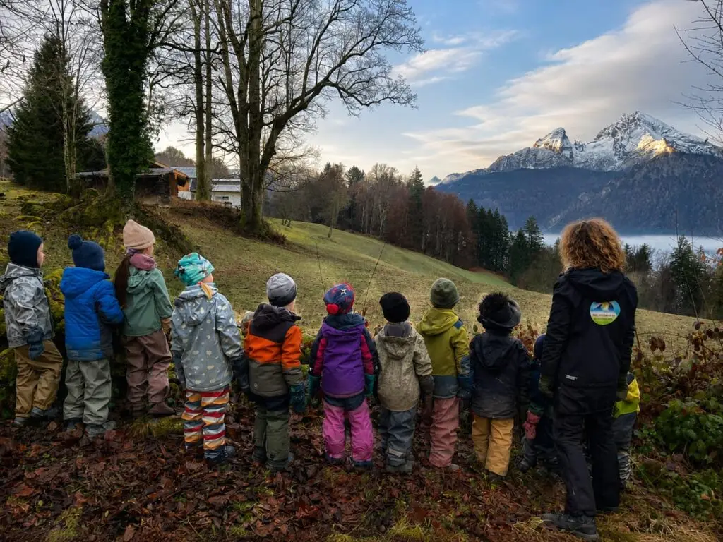 Eine Gruppe Kinder des Waldkindergartens Berchtesgaden steht in Matschkleidung am Waldrand und blickt auf den verschneiten Watzmann.